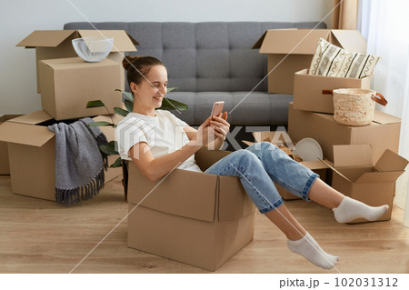 Portrait of happy Caucasian dark haired woman wearing white t shirt and jeans sitting in a cardboard box, holding mobile phone and scrolling online, relocating in her new flat. Portrait of happy Caucasian dark haired woman wearing white t shirt and jeans sitting in a cardboard box, holding mobile phone and scrolling online, relocating in her new flat. 102031312