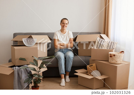 Indoor shot of beautiful positive woman wearing white casual T-shirt and jeans, sitting on sofa, surrounded with boxes during relocating, posing with folded hands, looking at camera. Indoor shot of beautiful positive woman wearing white casual T-shirt and jeans, sitting on sofa, surrounded with boxes during relocating, posing with folded hands, looking at camera. 102031326