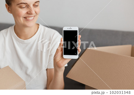 Satisfied woman wearing white t-shirt sitting on sofa, posing in a new apartment after relocation, showing cell phone with blank black screen for promotion of real estate and delivery service. 102031348
