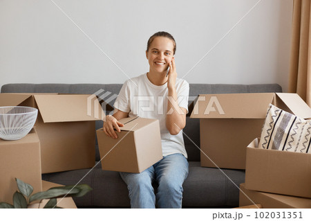 Indoor shot of attractive smiling woman wearing white shirt and jeans sitting on sofa, holding cardboard box with personal things and talking via mobile phone, looking at camera with smile. 102031351