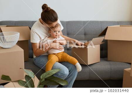 Portrait of woman with bun hairstyle wearing white t shirt sitting on sofa and holding her infant baby, unpacking boxes together with her daughter, young family moving into a new apartment. 102031440
