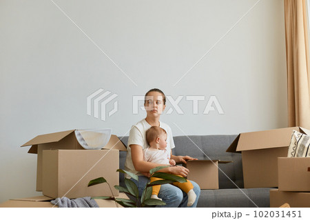 Portrait of young adult woman wearing white t shirt sitting on cough, posing infant baby, moving into a new apartment, looking away with puzzled and confused expression, thinking about interior. Portrait of young adult woman wearing white t shirt sitting on cough, posing infant baby, moving into a new apartment, looking away with puzzled and confused expression, thinking about interior. 102031451