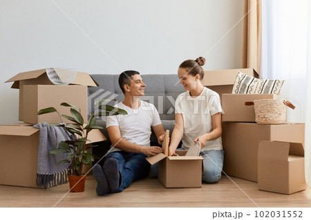 Caucasian family wearing casual attire relocating in new apartment, sitting on the floor near sofa among carton boxes, couple buying a house together, move in new flat, unpacking personal piles. Caucasian family wearing casual attire relocating in new apartment, sitting on the floor near sofa among carton boxes, couple buying a house together, move in new flat, unpacking personal piles. 102031552