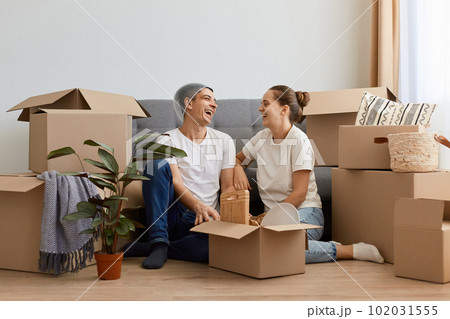 Happy satisfied newlyweds moved in new apartment, sitting on the floor and having fun together, laughing happily, husband with basket on head, relocated in new home. 102031555