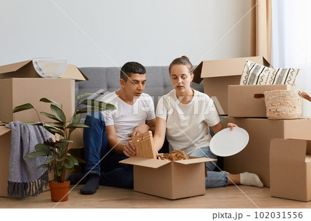 Indoor shot of shocked couple relocated in a new apartment and unpacking cardboard boxes with personal belongings, being surprised and sad on broken plates. 102031556