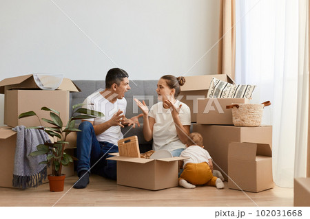 Indoor shot of sad aggressive couple having argument while moving to a new house, sitting on floor near sofa and screaming, quarelling during relocation in a new apartment. 102031668