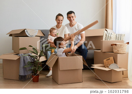 Horizontal shot of satisfied family unpacking moving box at their new house and having fun together, little girl sitting in carton box and looking in handmade spyglass. Horizontal shot of satisfied family unpacking moving box at their new house and having fun together, little girl sitting in carton box and looking in handmade spyglass. 102031798