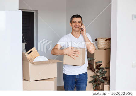 Horizontal shot of satisfied brunette man wearing white t shirt standing with cardboard box in hands, and talking on smart phone, looking at camera with positive facial expression. Horizontal shot of satisfied brunette man wearing white t shirt standing with cardboard box in hands, and talking on smart phone, looking at camera with positive facial expression. 102032050