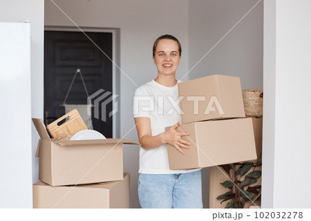 Indoor shot of happy smiling woman wearing white casual style T-shirt and jeans standing with cardboard boxes with belongings, looking at camera with positive expression, being glad to relocate. Indoor shot of happy smiling woman wearing white casual style T-shirt and jeans standing with cardboard boxes with belongings, looking at camera with positive expression, being glad to relocate. 102032278