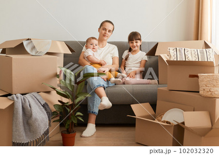 Indoor shot of Caucasian woman sitting on sofa with her daughters, looking at camera, unpacking packages with belongings, buying and moving to a new flat. Indoor shot of Caucasian woman sitting on sofa with her daughters, looking at camera, unpacking packages with belongings, buying and moving to a new flat. 102032320