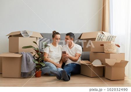 Indoor shot of young couple in love moving in a new apartment, sitting on the floor near, planning to redecorate their new home, man holding smart phone, showing ideas to his wife. Indoor shot of young couple in love moving in a new apartment, sitting on the floor near, planning to redecorate their new home, man holding smart phone, showing ideas to his wife. 102032490