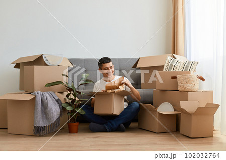 Horizontal shot of brunette positive man wearing white t shirt sitting on floor near sofa surrounded with cardboard boxes, upacking his personal piles after moving in. Horizontal shot of brunette positive man wearing white t shirt sitting on floor near sofa surrounded with cardboard boxes, upacking his personal piles after moving in. 102032764