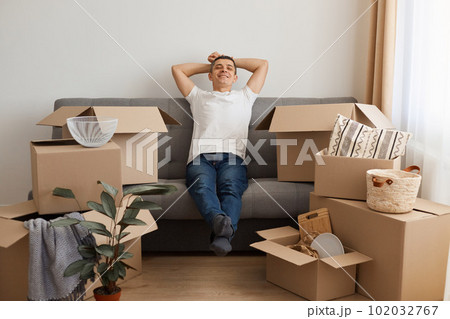 Happy satisfied relaxed man wearing white T-shirt and jeans sitting on sofa surrounded with pile of packages, looking at camera with smile, keeping hands behind head, being glad to move to a new flat. 102032767