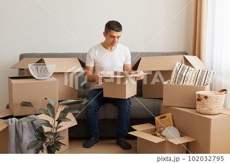 Sad man wearing white T-shirt and jeans sitting on sofa surrounded with cardboard boxes, posing with pout lips and sorrow expression, looking inside package, upset of broken things after moving. 102032795