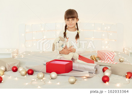 Dark haired little girl with pigtails wearing white sweater sitting in light bedroom on bed, holding soft toy dog in hands, looking at camera with charming positive facial expression. Dark haired little girl with pigtails wearing white sweater sitting in light bedroom on bed, holding soft toy dog in hands, looking at camera with charming positive facial expression. 102033364