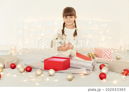 Good looking female girl wearing white sweater holding soft toy while sitting on bed with Christmas decoration and garland, kid with pigtails receives long awaited present. 102033365