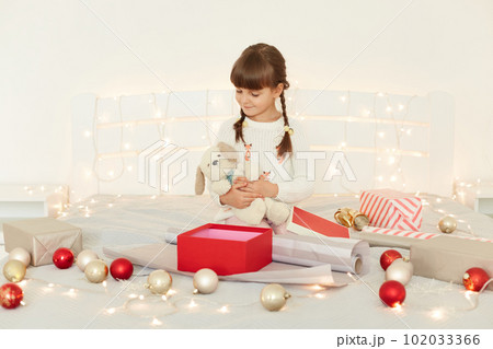 Charming little girl wearing white sweater embracing soft toy dog, sitting on bed with Christmas decoration and garland, looking at her present, celebrating New year eve. Charming little girl wearing white sweater embracing soft toy dog, sitting on bed with Christmas decoration and garland, looking at her present, celebrating New year eve. 102033366