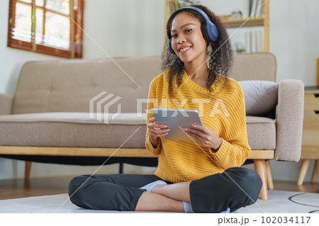 Portrait of an African American with headphones and tablet smiling happily listen music while relaxing on the sofa at home. Portrait of an African American with headphones and tablet smiling happily listen music while relaxing on the sofa at home. 102034117