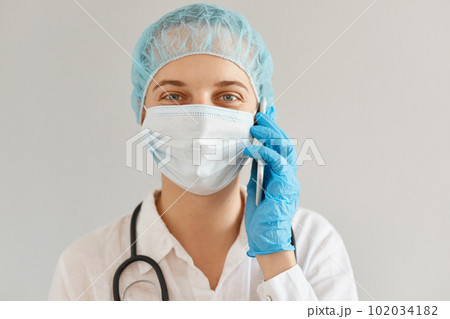 Smiling positive female doctor wearing medical uniform, cap and surgical mask talking phone with patient, looking at camera with optimistic facial expression. 102034182