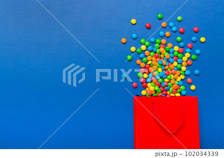shopping paper gift bag in corner full of assorted traditional candies falling out on colored background with copy space. Happy Holidays sale concept shopping paper gift bag in corner full of assorted traditional candies falling out on colored background with copy space. Happy Holidays sale concept 102034339
