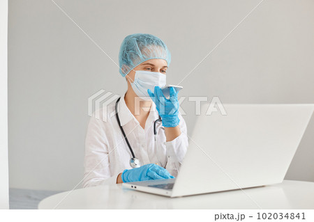 Portrait of serious confident female doctor wearing medical cap, surgical mask, rubber gloves and gown, using notebook for working and cell phone for recording voice message. 102034841