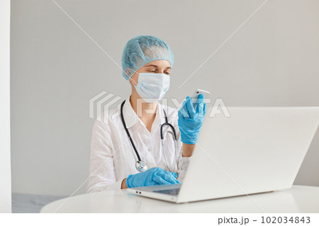 Confident woman doctor sitting at table and working on laptop computer, holding mobile phone in hands for checking e mails, wearing medical cap, surgical mask, rubber gloves and gown. Confident woman doctor sitting at table and working on laptop computer, holding mobile phone in hands for checking e mails, wearing medical cap, surgical mask, rubber gloves and gown. 102034843
