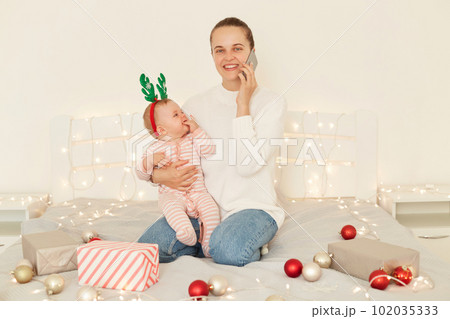 Attractive young adult happy woman with positive expression sitting on bed with baby girl in arms and talking phone, congratulating somebody with Christmas and Happy new year. Attractive young adult happy woman with positive expression sitting on bed with baby girl in arms and talking phone, congratulating somebody with Christmas and Happy new year. 102035333