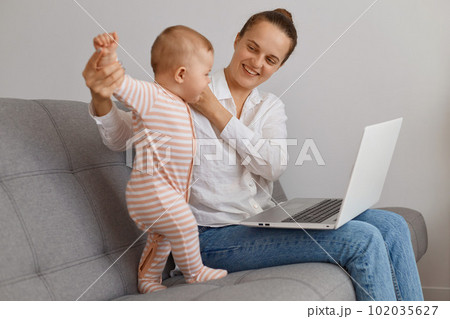 Delighted smiling dark haired woman spending time with toddler daughter and working on notebook, sitting on sofa in living room, expressing positive emotions. Delighted smiling dark haired woman spending time with toddler daughter and working on notebook, sitting on sofa in living room, expressing positive emotions. 102035627