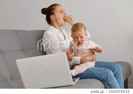Side view portrait of tired woman with hair bun in white shirt and jeans sitting on cough with daughter, yawning, holding crying baby, covering mouth with hand, having rest after working on computer. Side view portrait of tired woman with hair bun in white shirt and jeans sitting on cough with daughter, yawning, holding crying baby, covering mouth with hand, having rest after working on computer. 102035635