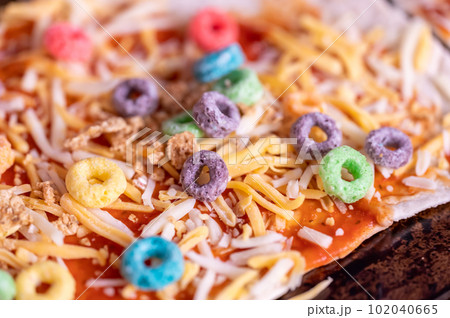 Pizza made by a child with cereal, cheese, and sauce. 102040665