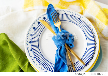 Serving design. Easter serving concept. Gold cutlery is tied with a blue napkin, on which the plates are on a green-yellow linen napkin. Minimalistic design. Serving design. Easter serving concept. Gold cutlery is tied with a blue napkin, on which the plates are on a green-yellow linen napkin. Minimalistic design. 102040722