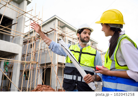 Young attractive construction man and woman in vests with helmets working on the under-construction building site. Home building project. Engineer foreman discusses with a coworker at workplace. 102045258