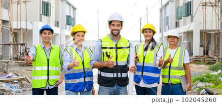 Group of happy contractors, engineers and formats in safety vests with helmets showing thumb up while standing on the under-construction building site. teamwork concept. 102045260