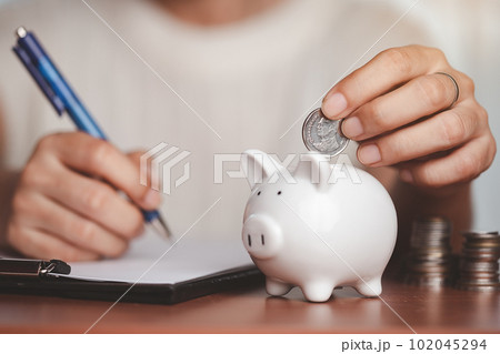 Hand saving a coin into piggy bank, planning and writing information on paper with blurred coins and money on the wooden 102045294
