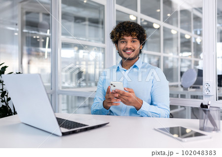 A young Latin American businessman sits in the office at a desk and uses a mobile phone, writes a message, searches for information on the Internet, checks social networks. Smiling at the camera. A young Latin American businessman sits in the office at a desk and uses a mobile phone, writes a message, searches for information on the Internet, checks social networks. Smiling at the camera. 102048383