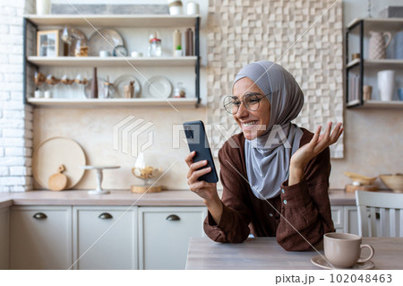 Close-up photo. Smiling Muslim young woman in hijab standing in kitchen at home, leaning on table and talking on mobile phone video call. 102048463