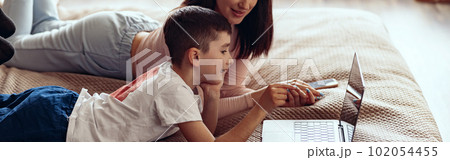 Small Caucasian cute boy with beautiful mother lying together on bed with laptop. 102054455