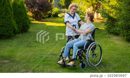 An elderly female nurse walks with a middle-aged woman in a wheelchair in the park. An elderly female nurse walks with a middle-aged woman in a wheelchair in the park. 102065697