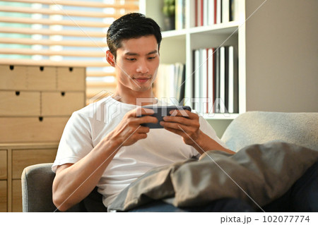 Happy millennial man playing video games on smart phone while resting on couch.Technology, entertainment and leisure activity Happy millennial man playing video games on smart phone while resting on couch.Technology, entertainment and leisure activity 102077774
