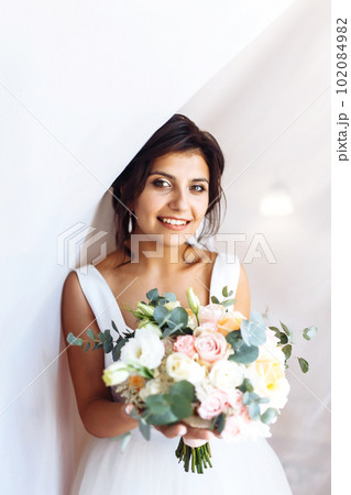 Young bride in white dress with a bouquet of flowers posing under curtain. Morning of the bride. Young bride in white dress with a bouquet of flowers posing under curtain. Morning of the bride. 102084982