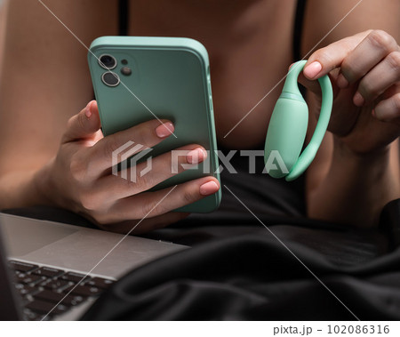 A woman lies on a bed and synchronizes the kegel machine with a smartphone. A woman lies on a bed and synchronizes the kegel machine with a smartphone. 102086316