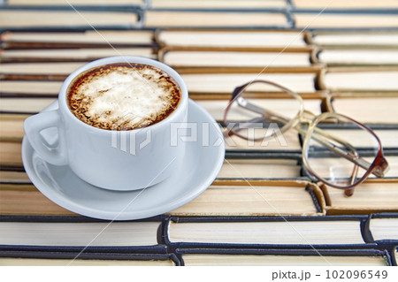 A white cup of fragrant cappuccino coffee and reading glasses on the background of worn old books folded upside down 102096549