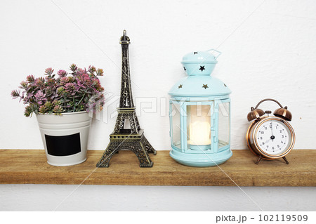 Eiffel tower statue, artificial plant, vintage alarm clock and blue lantern with LED candle light on wooden shelves Eiffel tower statue, artificial plant, vintage alarm clock and blue lantern with LED candle light on wooden shelves 102119509