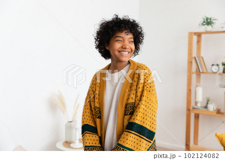 Beautiful african american girl with afro hairstyle smiling at home indoor. Young african woman with curly hair laughing in living room. Freedom happiness carefree happy people concept 102124082