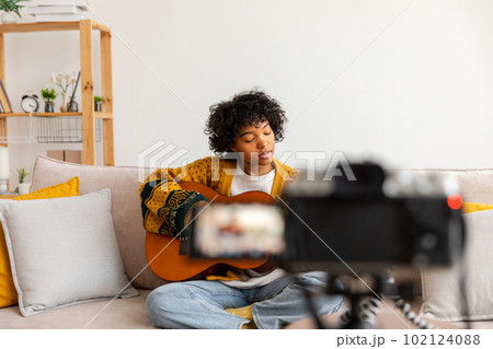 Blogger guitarist. African american girl blogger playing guitar talking to webcam recording vlog. Social media influencer woman streaming at home indoors. Music content creator broadcast tutorial 102124088