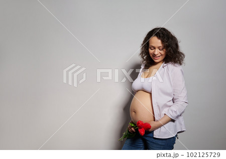 Middle aged beautiful pregnant woman expecting baby, holding tulips near her belly, posing with happy and cool smile on face, over isolated white background. Copy advertising space. Pregnancy fashion. Middle aged beautiful pregnant woman expecting baby, holding tulips near her belly, posing with happy and cool smile on face, over isolated white background. Copy advertising space. Pregnancy fashion. 102125729