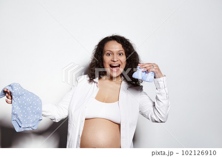 Overjoyed emotional multi ethnic curly haired pregnant woman holding baby clothes, blue bodysuit and booties for newborn boy, smiles looking at camera, posing bare belly over isolated white background Overjoyed emotional multi ethnic curly haired pregnant woman holding baby clothes, blue bodysuit and booties for newborn boy, smiles looking at camera, posing bare belly over isolated white background 102126910