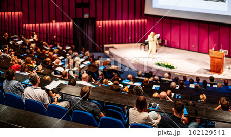 Business and entrepreneurship symposium. Female speaker giving a talk at business meeting. Audience in conference hall. Rear view of unrecognized participant in audience. 102129541