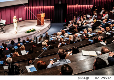 Business and entrepreneurship symposium. Female speaker giving a talk at business meeting. Audience in conference hall. Rear view of unrecognized participant in audience. 102129542