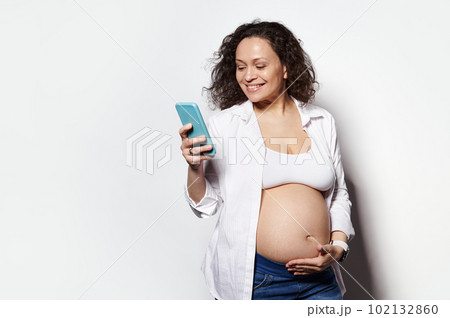 Curly haired multi ethnic brunette, cheerful happy woman in 30 weeks of pregnancy smiling while recording baby kicks on mobile application in her smartphone, isolated white background. Copy ad space Curly haired multi ethnic brunette, cheerful happy woman in 30 weeks of pregnancy smiling while recording baby kicks on mobile application in her smartphone, isolated white background. Copy ad space 102132860